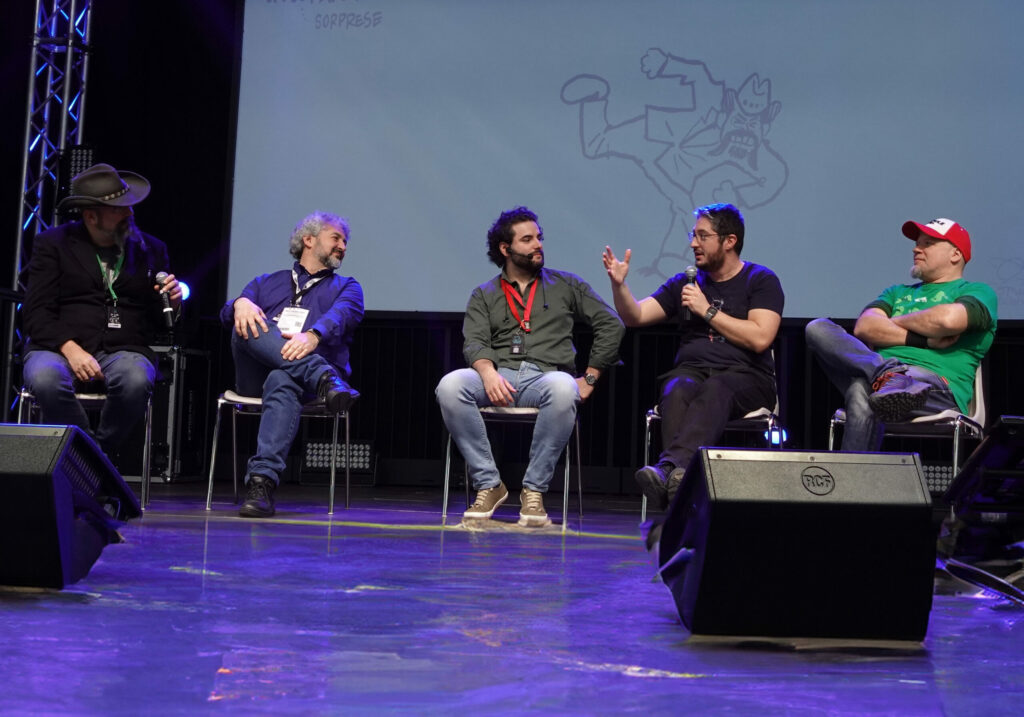 Quattro uomini sul palco stanno disquisendo. Alle loro spalle un monitor gigante dove ci sono alcune scritte, tra cui il nome del match "Calci&Sputi"