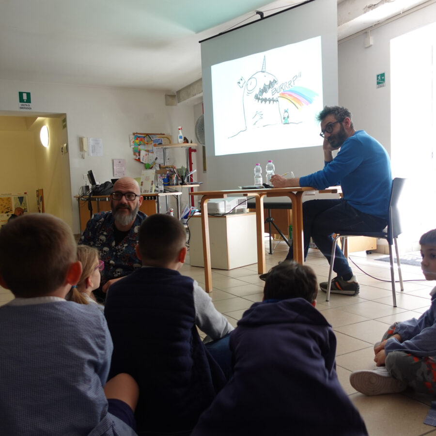 Bambini visti di spalle parlano a Diego Cajelli (pelato, con barba e occhiali scuri). A lato, Andrea Scoppetta ascolta e aspetta a disegnare davanti al suo iPad. Sullo sfondo la proiezione dei disegni che sta facendo.