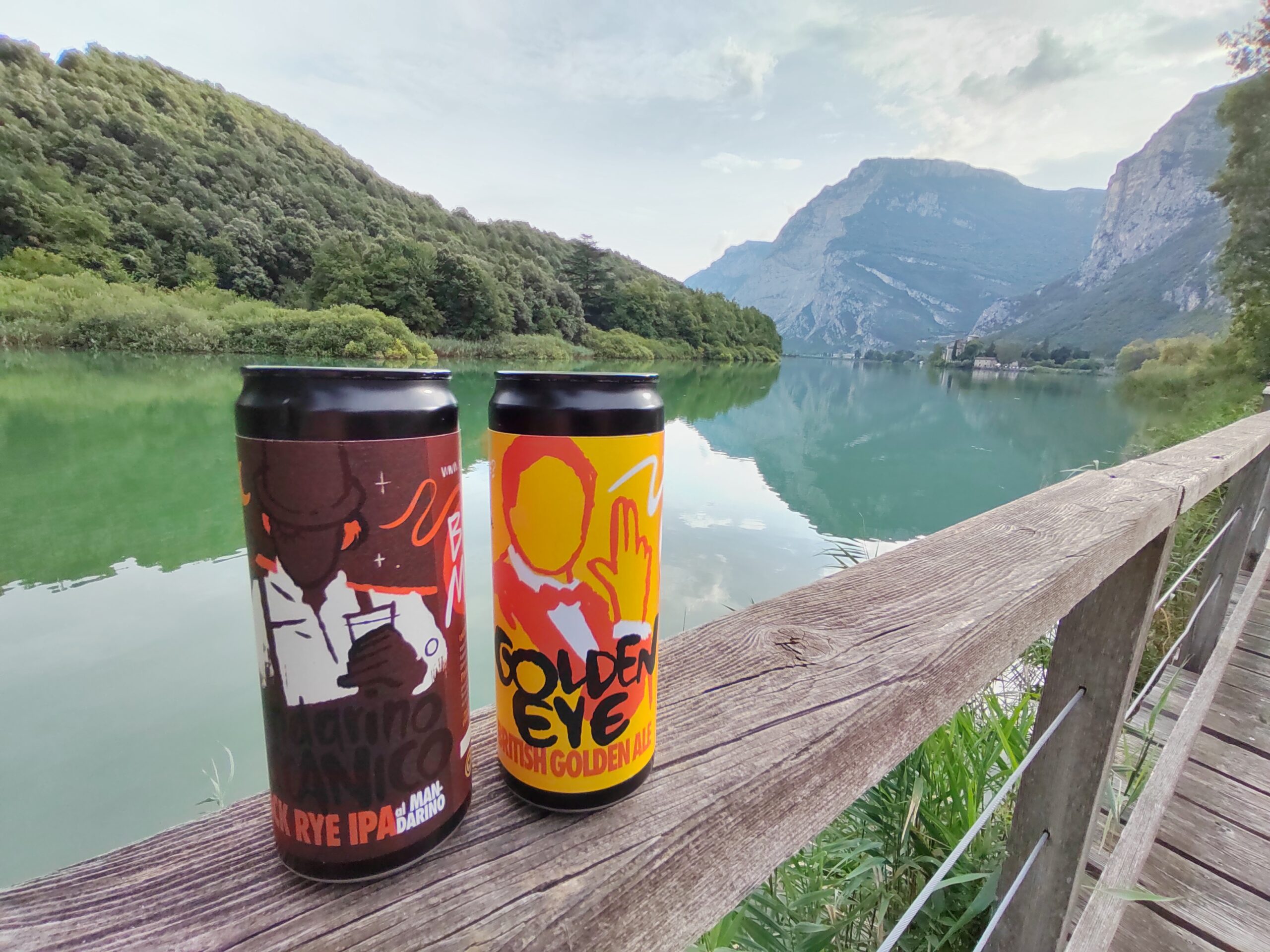 Due lattine colorate sono appoggiate alla ringhiera di legno di un sentiero di montagna. Il percorso dà su un lago dalle acque azzurro-verde. Sullo sfondo si vedono le montagne.